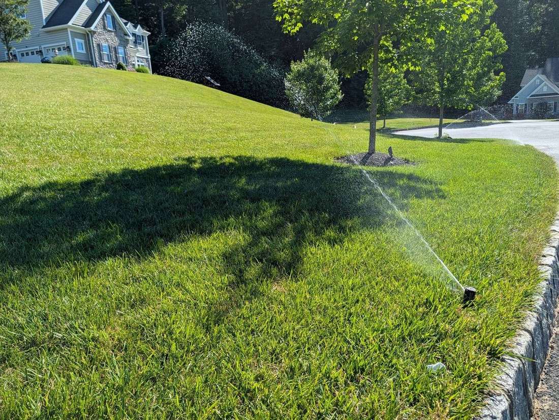 A pop-up lawn sprinkler spraying an arc of water across a lush green front lawn in front of a stone-front colonial home in Newtown Square, PA
